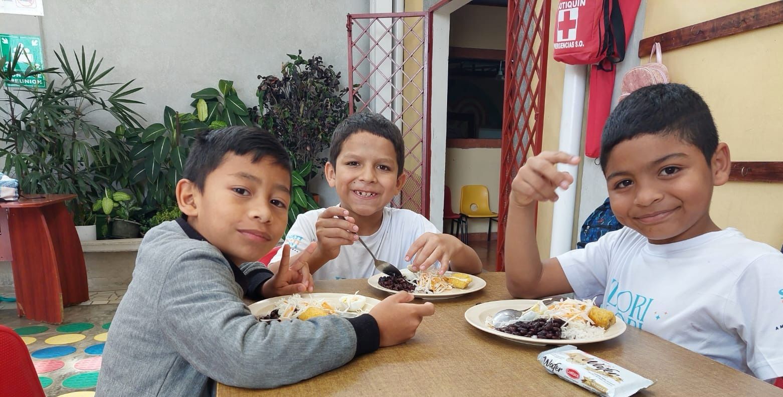 Tres niños están sentados ante una mesa mientras sonríen y comen un plato de comida.