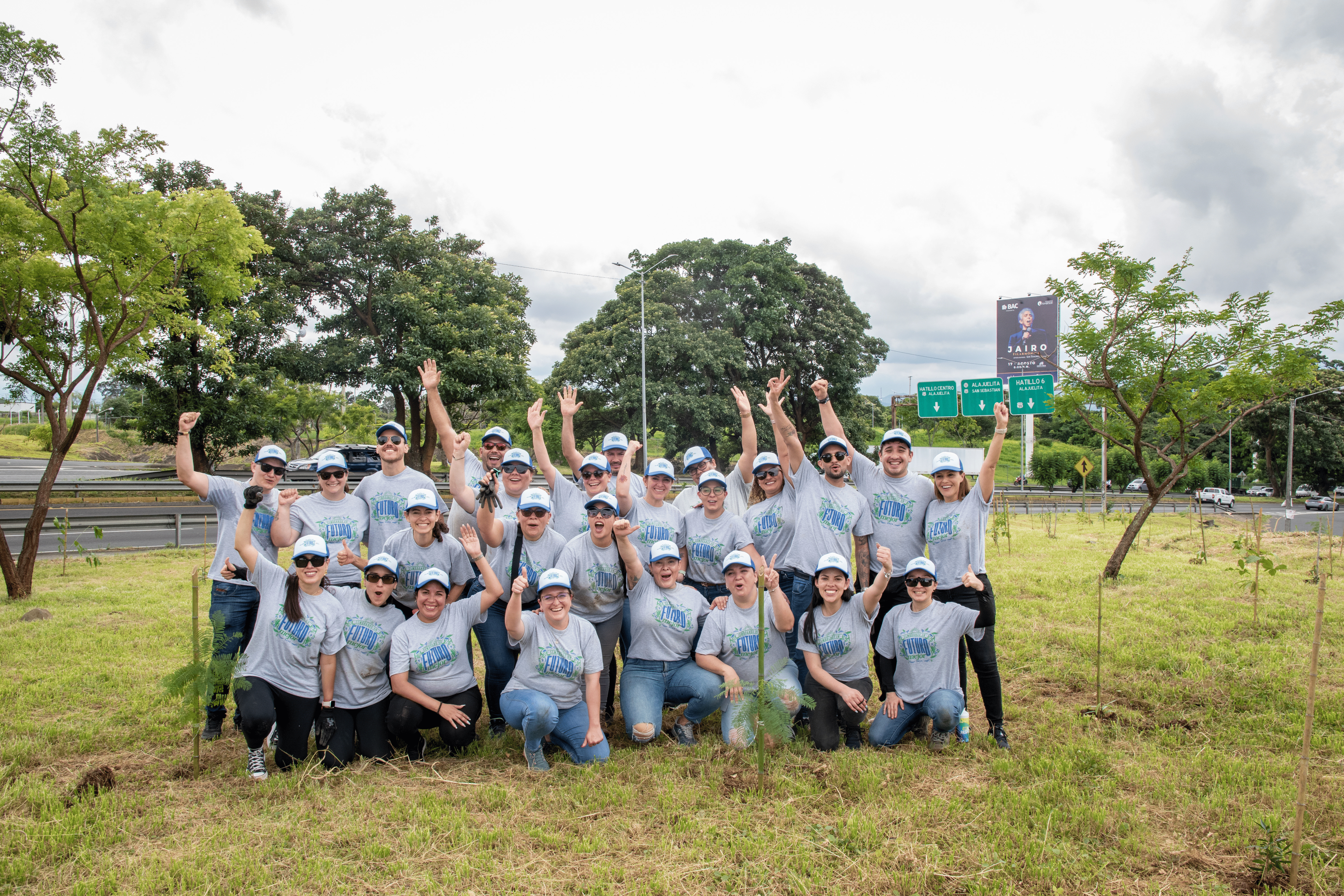 Un grupo de voluntarios vestidos con camisas grises posa en un terreno verde, en medio de pequeños árboles recién plantados.