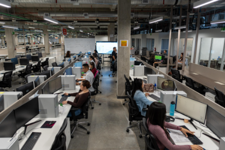 Amplio espacio de oficina con varias estaciones de trabajo en filas, donde numerosos empleados están sentados frente a sus computadoras. El diseño es de estilo abierto, con divisiones bajas entre los escritorios. Al fondo, una persona parece dar una presentación frente a una pantalla grande, y se observa un ambiente moderno, con columnas de concreto y tuberías expuestas en el techo. La oficina tiene un ambiente de actividad y colaboración.