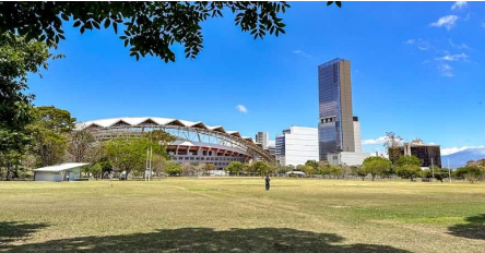 Amplio parque con áreas de césped y algunos árboles en primer plano, que ofrece una vista hacia el Estadio Nacional y un edificio alto y moderno en el horizonte. El cielo está despejado y azul, y se puede ver la estructura distintiva del estadio en contraste con el edificio de gran altura y otras edificaciones en la zona urbana. Un par de personas disfrutan del espacio abierto bajo la luz del sol.