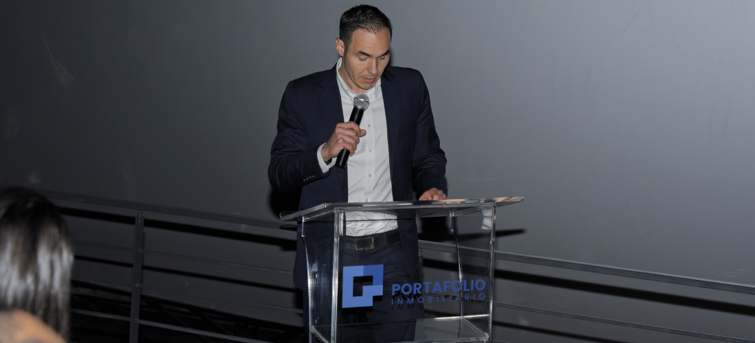 "Alfredo Volio director ejecutivo de la empresa viste de traje oscuro, camisa blanca y corbata se encuentra de pie detrás de un podio transparente con el logotipo de Portafolio Inmobiliario. Está sosteniendo un micrófono mientras parece dirigirse a una audiencia en un evento formal."